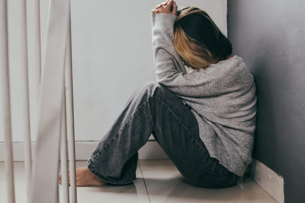 Person sits on a tiled floor in a corner, knees drawn up, hands resting on their head, wearing a gray knit sweater and dark jeans, conveying distress or sadness.